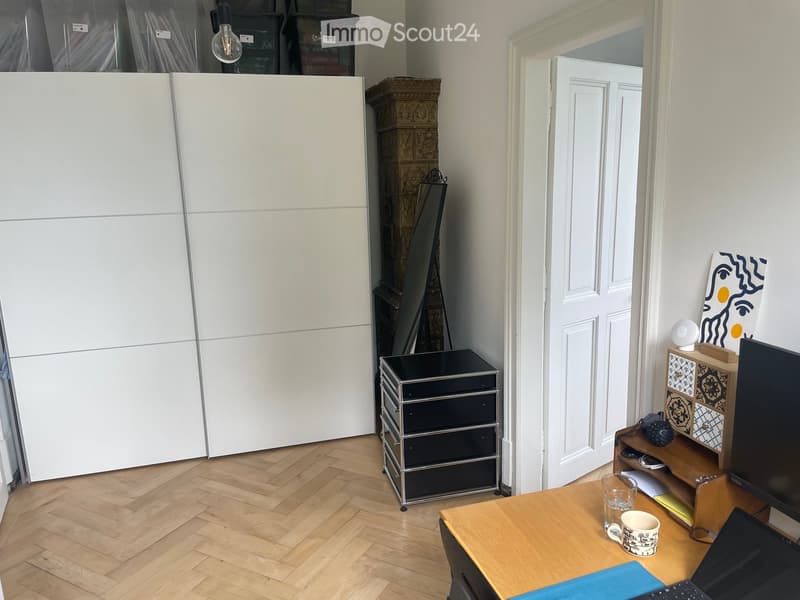 A furnished room with a wooden floor, a large white cabinet, a wooden table with a laptop, a cup, and a screen, along with a small black cabinet and a wooden shelf with decorative items.