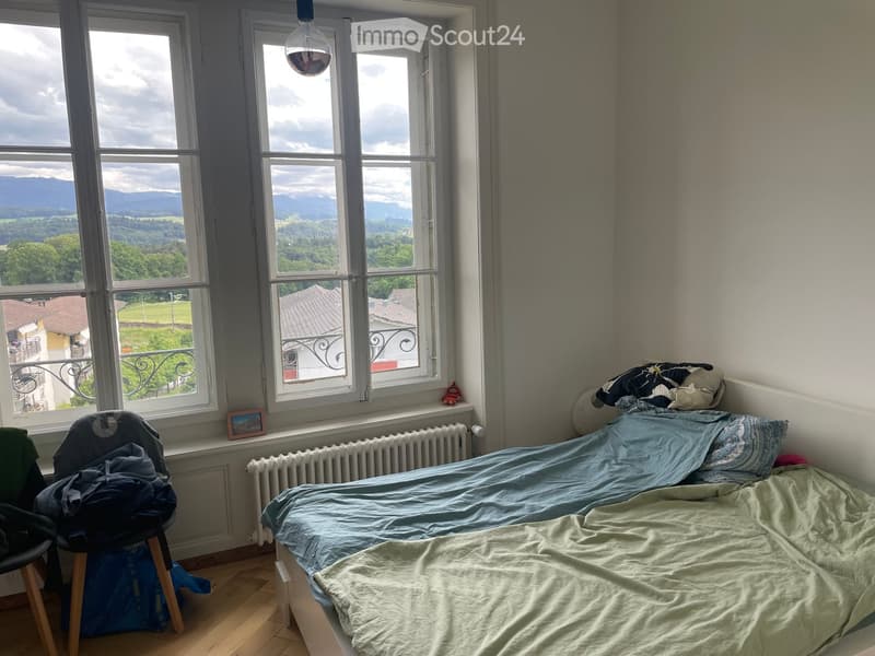 Bedroom with a single bed, wooden floor, large windows offering a scenic view, a chair with clothes, and a radiator.