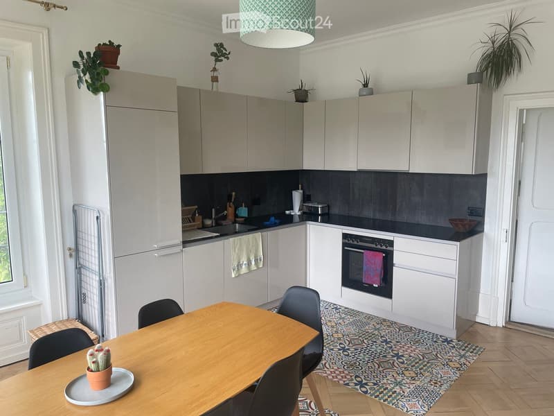 Modern kitchen with white cabinets, dark countertops, and a wooden dining table with chairs. Features include a refrigerator, oven, sink, and various kitchen appliances.
