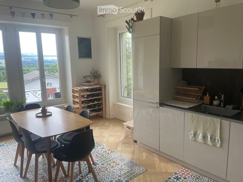 A modern, well-lit kitchen and dining area with a wooden dining table, chairs, a wine rack, and sleek kitchen cabinets. The space features a view of a scenic landscape through large windows.