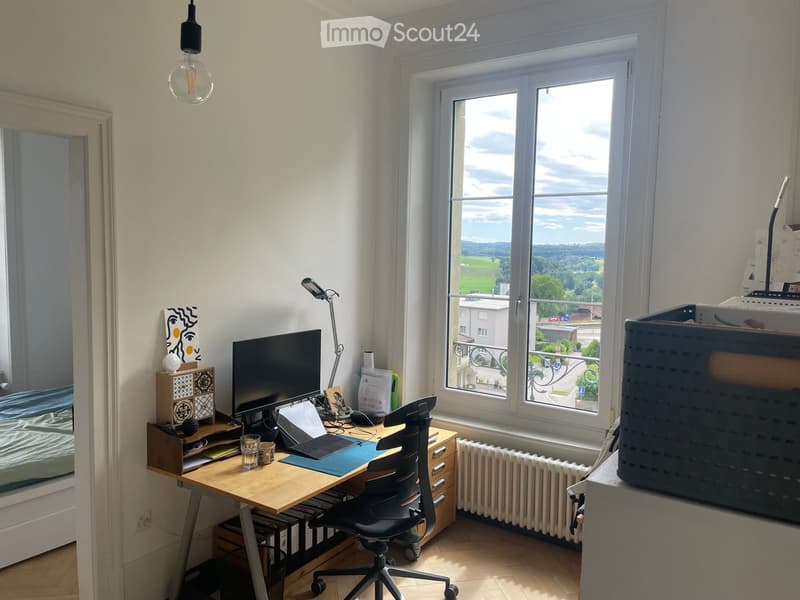 A well-lit room with a wooden desk, ergonomic chair, laptop, monitor, and various office supplies. Large windows offer a scenic view of the landscape.