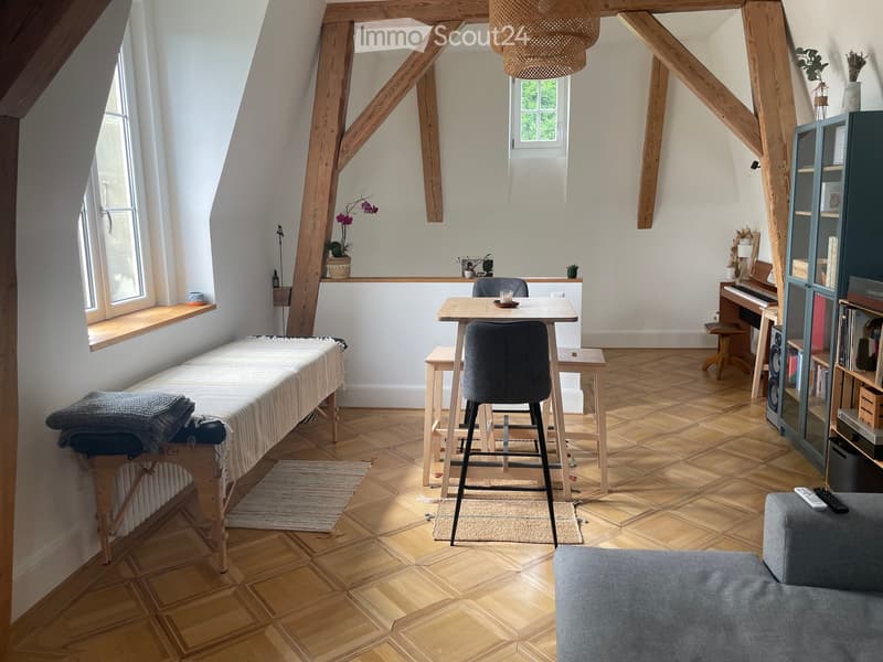 Bright, airy room with wooden beams, wooden floor, large windows, sofa, piano, table, chairs, and decorative items.