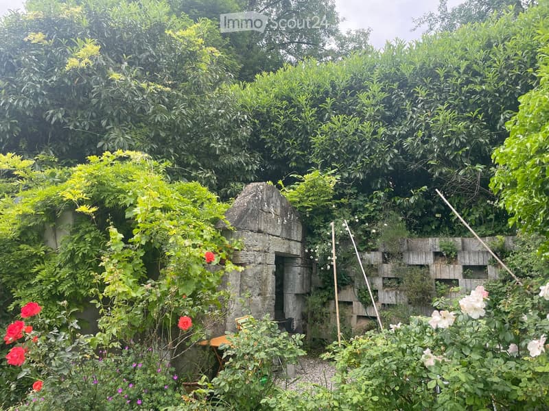 A lush garden with a stone well surrounded by dense greenery, roses, and climbing plants. A small wooden bench is visible near the well.