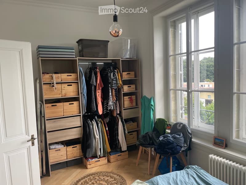 Well-lit bedroom with wooden floor, large window, wardrobe with hanging clothes and wooden drawers, stool with folded clothes, picture frame on windowsill, radiator, and wicker basket on floor.