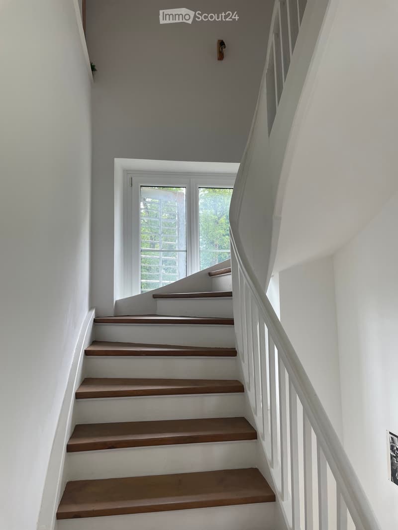 A modern staircase with wooden steps and white railings, leading to a bright, open landing area with a large window overlooking greenery.