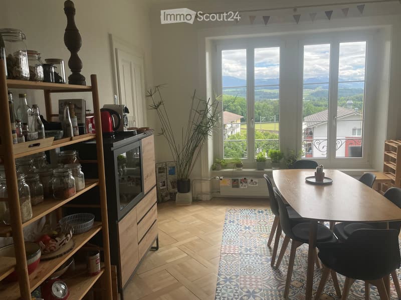 Bright kitchen with wooden flooring, large windows overlooking a scenic view, a dining table with chairs, shelves with jars and bottles, and a modern oven.