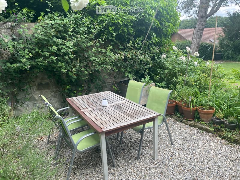 A small outdoor seating area with a wooden table and four green chairs, surrounded by potted plants and lush greenery.