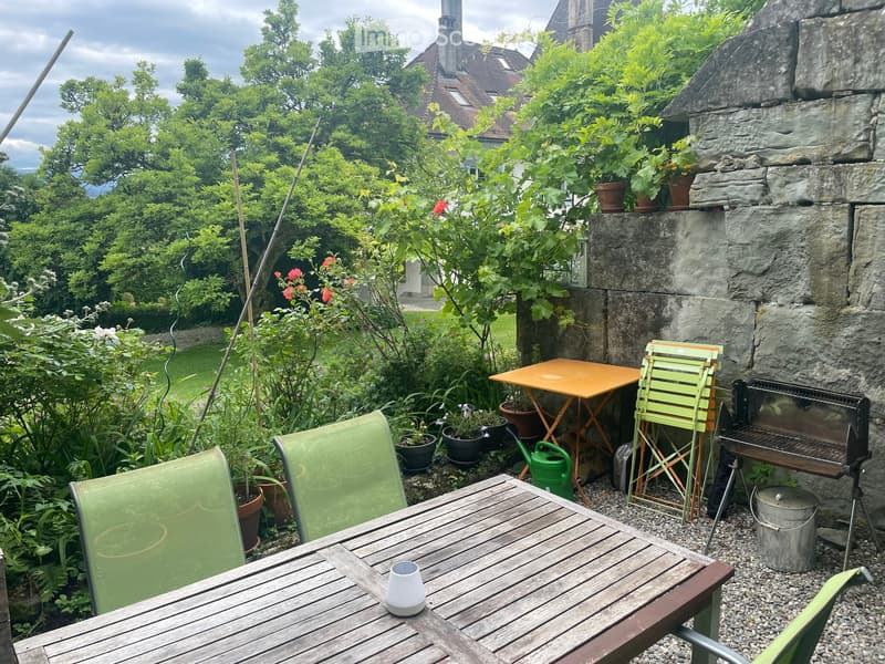 A small garden area with a wooden table, green chairs, a barbecue, potted plants, and a small orange table with a matching chair.