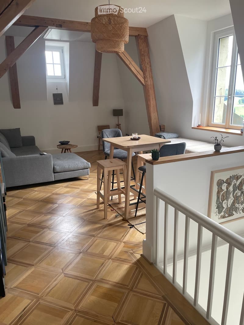A modern, open-concept living space with a grey sofa, ottoman, small table, and wooden stools. The room features exposed wooden beams, large windows, and a cozy, inviting atmosphere.