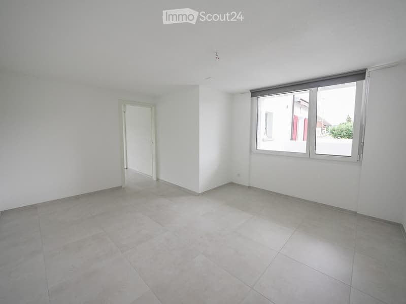 Empty room with white walls, tiled floor, and a single door leading to another space. Large windows with blinds allow natural light to enter.