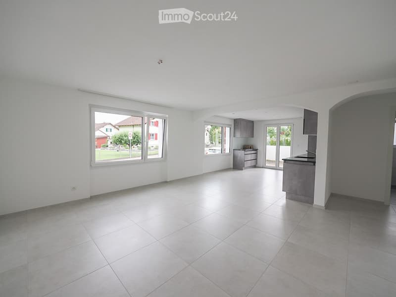 Empty room with tiled floor and white walls, featuring large windows and a small kitchen area with cabinets and a sink.