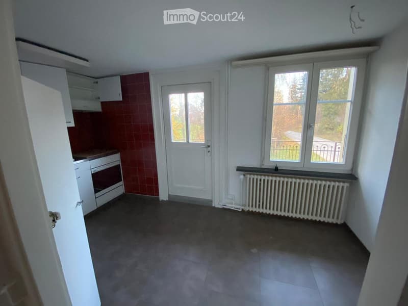 Empty kitchen with white cabinets, a white oven, a radiator, and a door leading outside. The floor is tiled, and the walls are painted white with red tiles in the backsplash area.