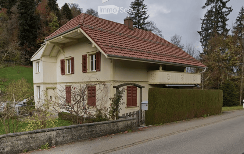 Two-story house with red-tiled roof, cream-colored walls, red shutters, and a small balcony on the upper floor.