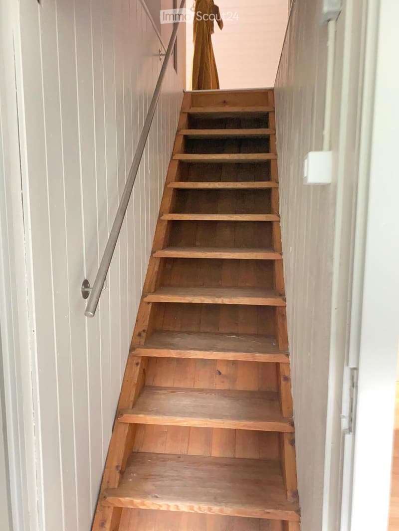 Wooden staircase with a metal handrail, light grey walls, and a partially visible door on the left side.