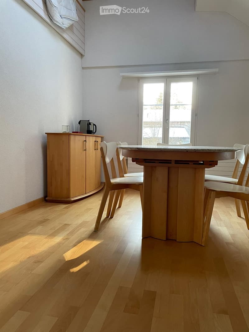 Bright dining area with wooden floor, round wooden table, four chairs, and a wooden cabinet with a coffee maker on top.