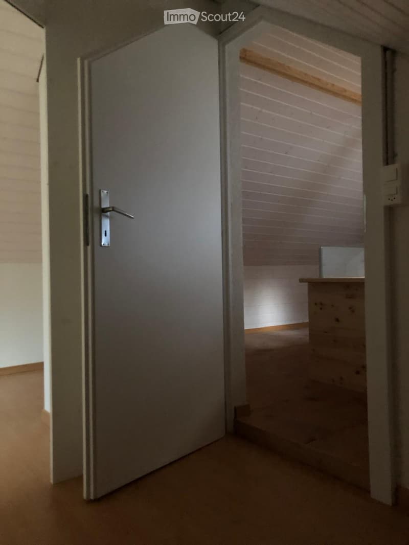 A narrow hallway with wooden walls and a closed door on the left, leading to a small room with a wooden bench and a small table.
