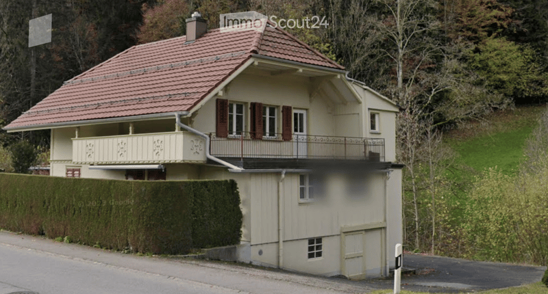 Two-story house with a red-tiled roof, beige exterior, and a balcony on the upper level. The house features multiple windows with shutters and a small garage attached to the lower level.
