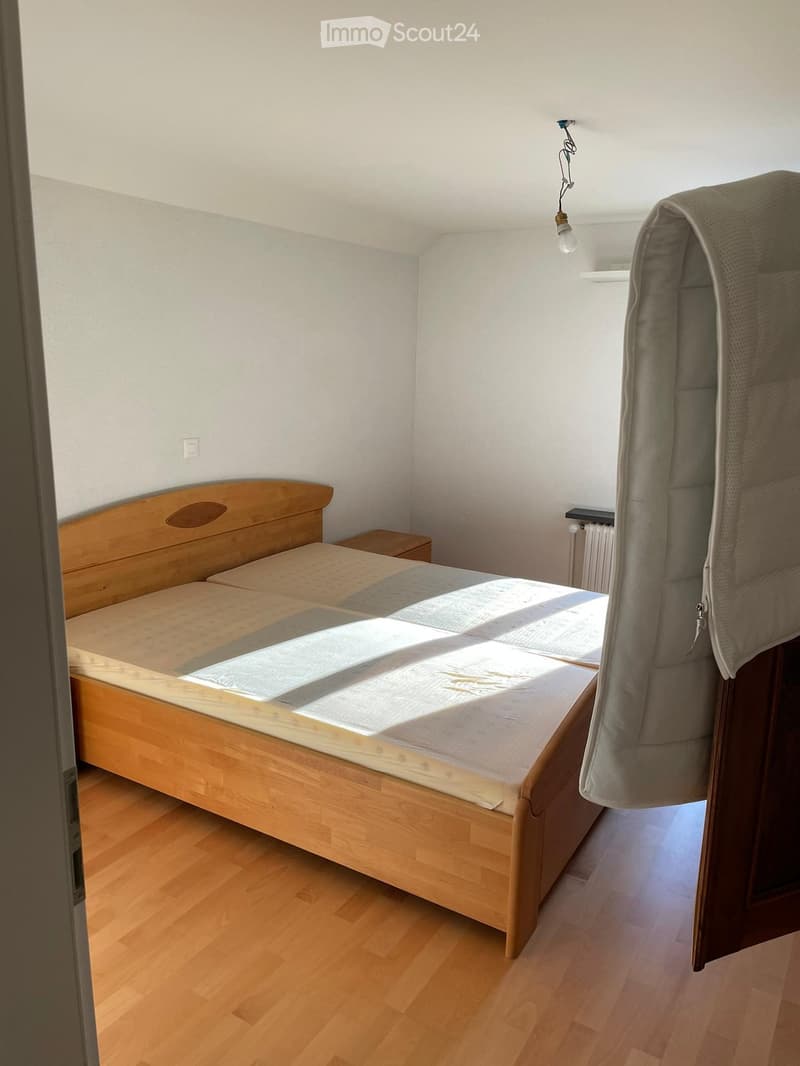Bright bedroom with wooden floor, double bed with mattress and bedding, nightstand, radiator, and hanging fabric on the right side.