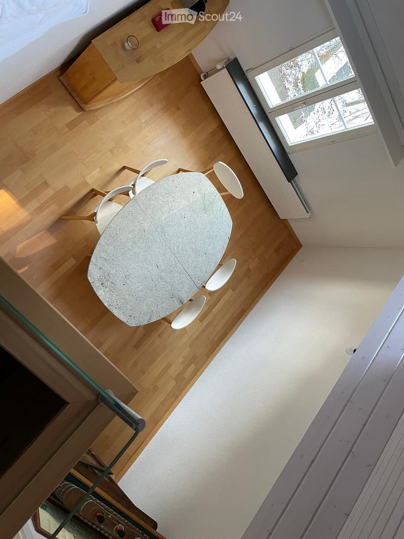 A modern, minimalist room with a round concrete table and four white chairs, wooden flooring, and a large window letting in natural light.
