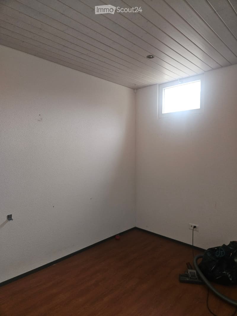 Empty room with wooden flooring, white walls, and a small window letting in natural light. There is a vacuum cleaner and some cables visible on the floor.
