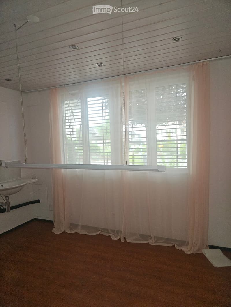Empty room with wooden flooring, white walls, and a window with blinds and curtains. A sink is visible on the left side.