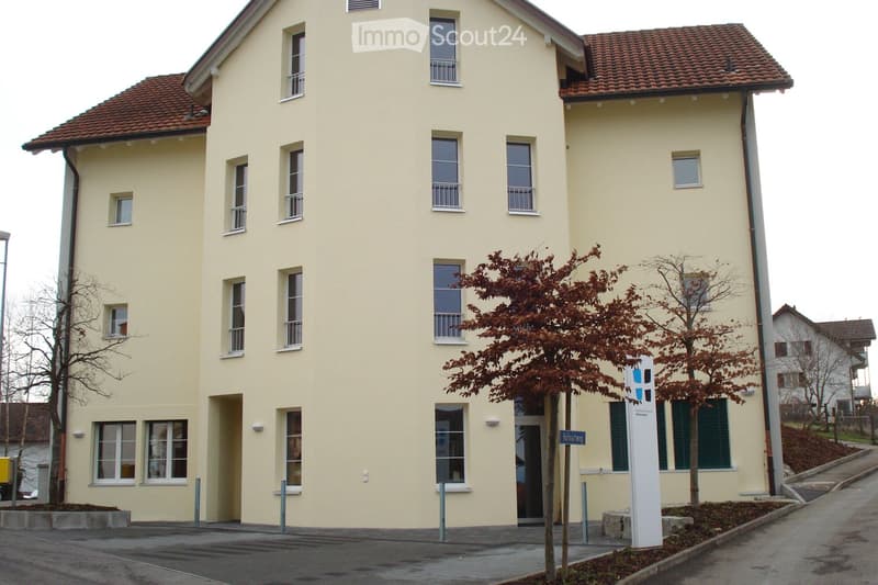 Four-story residential building with light yellow facade, multiple windows, and a red-tiled roof. Features a small tree in front and a street sign.