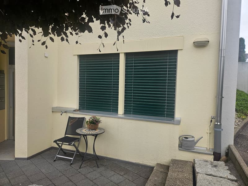 Balcony with a small round table, a folding chair, and a flower pot, adjacent to a window with green blinds.