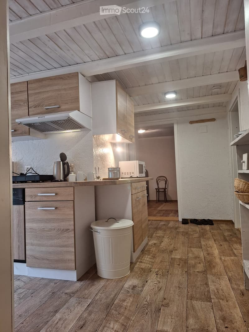 The kitchen features wooden cabinets, a white countertop, and a variety of appliances including a microwave, stove, and oven. The floor is made of light wooden planks, and there is a small chair near the entrance.