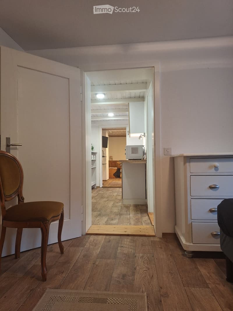 A narrow hallway with wooden flooring, leading to a kitchen area visible in the background. The hallway features a wooden chair on the left and a small cabinet on the right.