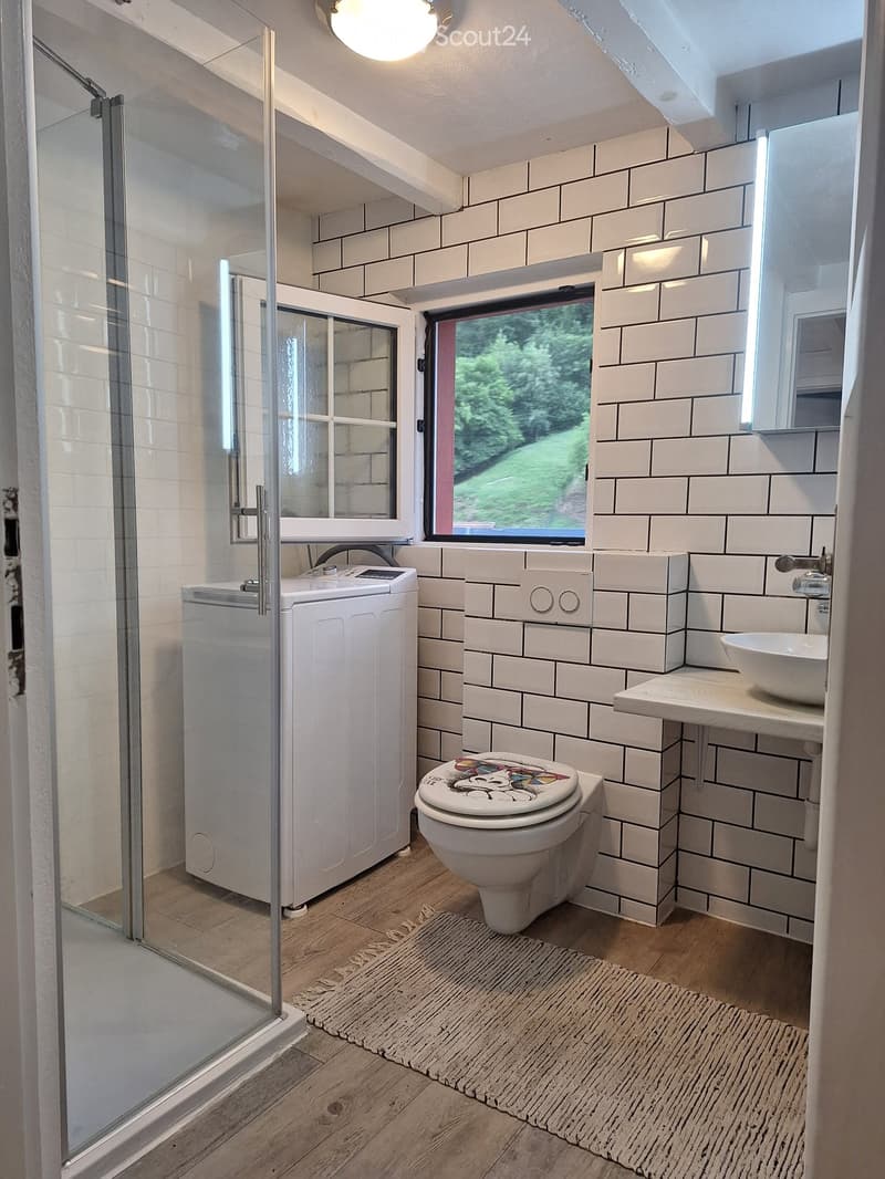 Modern bathroom with white tiled walls, wooden floor, toilet, sink, shower, and washing machine. Features a large window with a view of greenery.