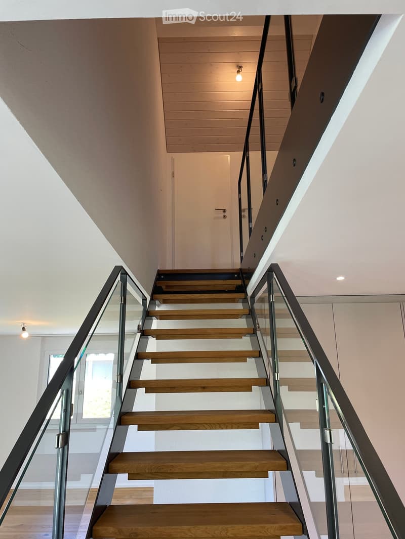 Modern staircase with wooden steps and glass railing, leading to an upper level with a visible door and window.