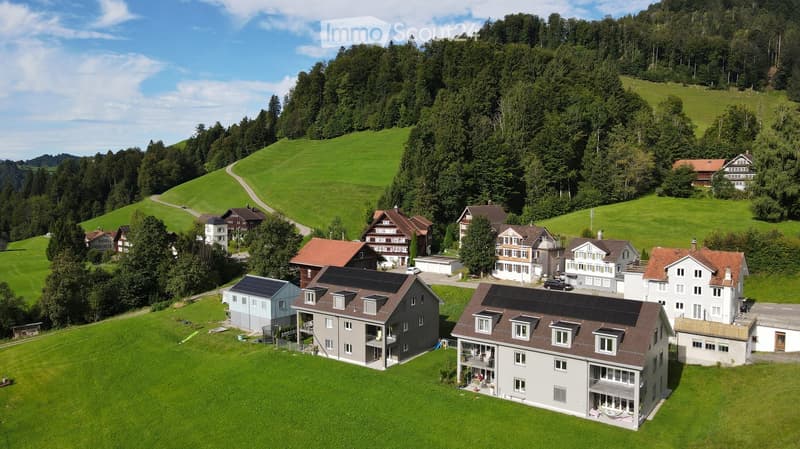 Aerial view of a residential area with multiple houses, surrounded by green hills and trees. Houses feature various architectural styles, some with solar panels on the roofs.