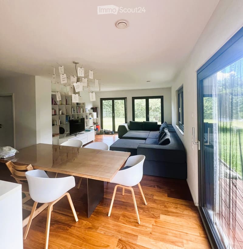 Modern living and dining area with a dark grey sectional sofa, a wooden dining table with four white chairs, a television on a white stand, and a bookshelf with books and decorative items.