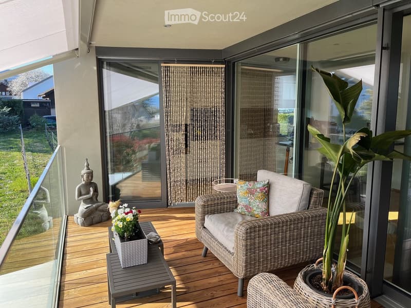 Balcony with wooden flooring, glass railing, wicker sofa, small table with flowers, large potted plant, and a decorative statue.