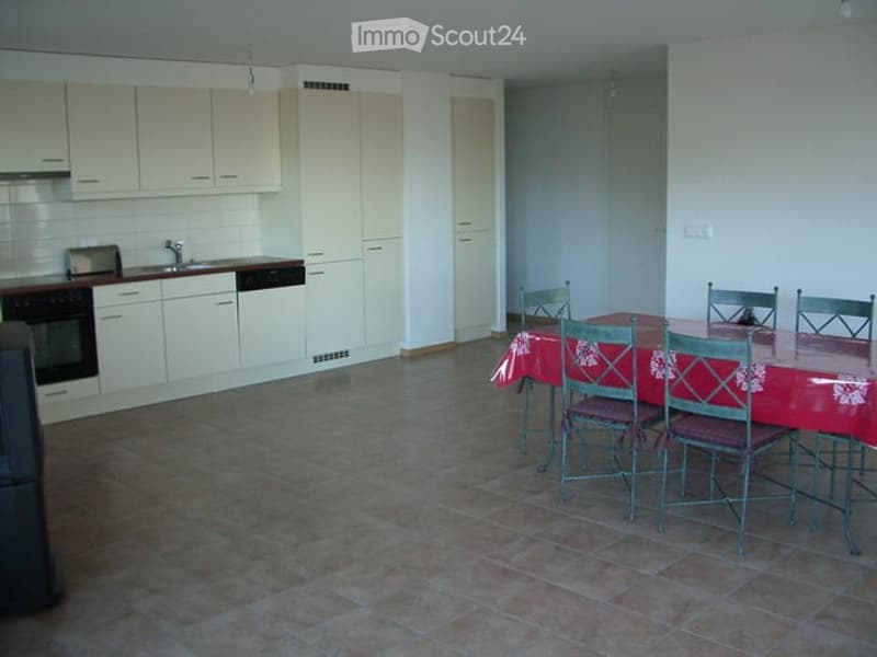 A modern kitchen with white cabinets, a sink, an oven, and a dishwasher. A dining area features a table with chairs and a red tablecloth.