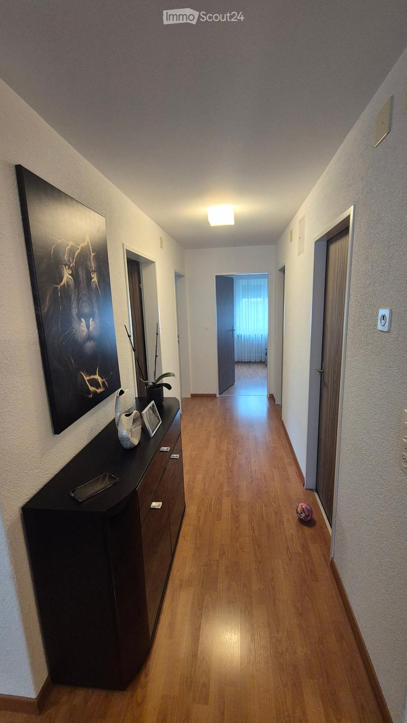 A well-lit hallway with wooden flooring, white walls, and several doors leading to different rooms. A small black cabinet with decorative items sits on the left side, and a colorful ball is placed near the right wall.