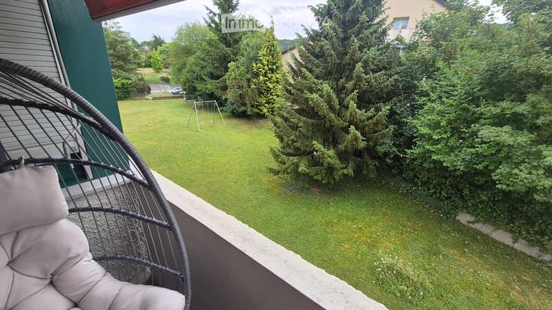 A well-maintained garden with green grass, trees, and a swing set visible in the background. A wicker hanging chair is placed on a concrete balcony.