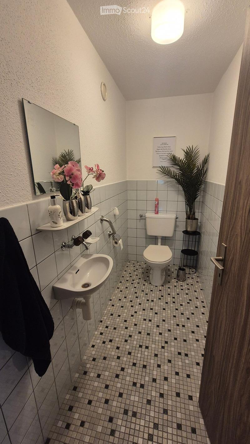 Modern bathroom with white tiled walls, black and white checkered floor, a toilet, a sink with a mirror, a towel, a plant, and a decorative flower vase.