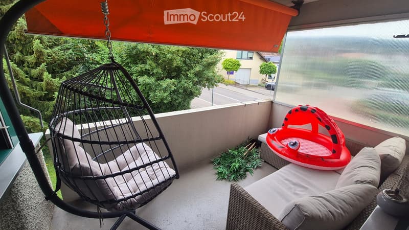 Balcony with a hanging egg chair, a wicker sofa, a red inflatable toy, and some potted plants.