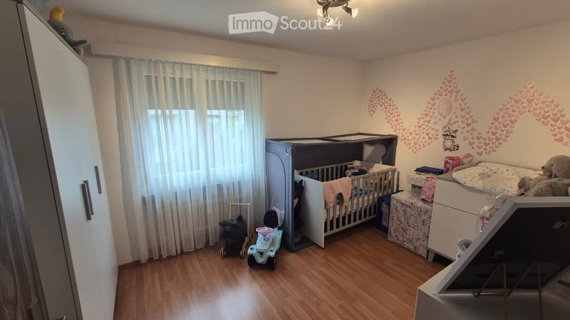 A well-lit children's bedroom with a crib, toy car, toy dog, and a wardrobe. The walls feature heart and animal decals, and the room has wooden flooring and white curtains.