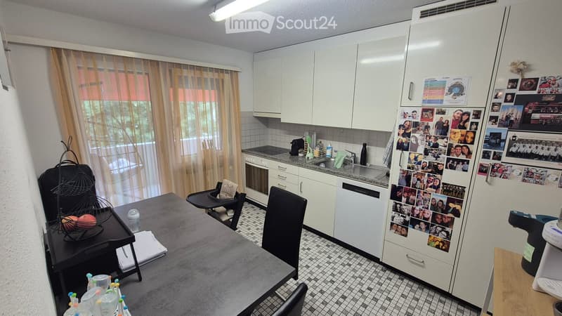 Modern kitchen with white cabinets, stainless steel appliances, and a large window with curtains. The kitchen features a refrigerator covered with photos, a sink, an oven, and a dining area with a table and chairs.