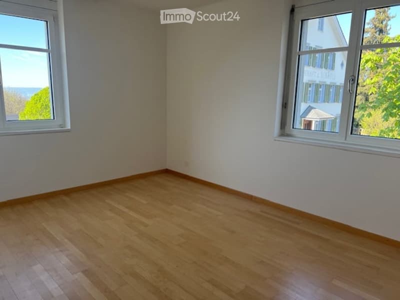 Empty room with wooden flooring and large windows offering a view of greenery and the sea.