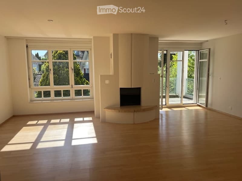 Bright, unfurnished living room with wooden flooring, large windows, fireplace, and sliding glass doors leading to a balcony.
