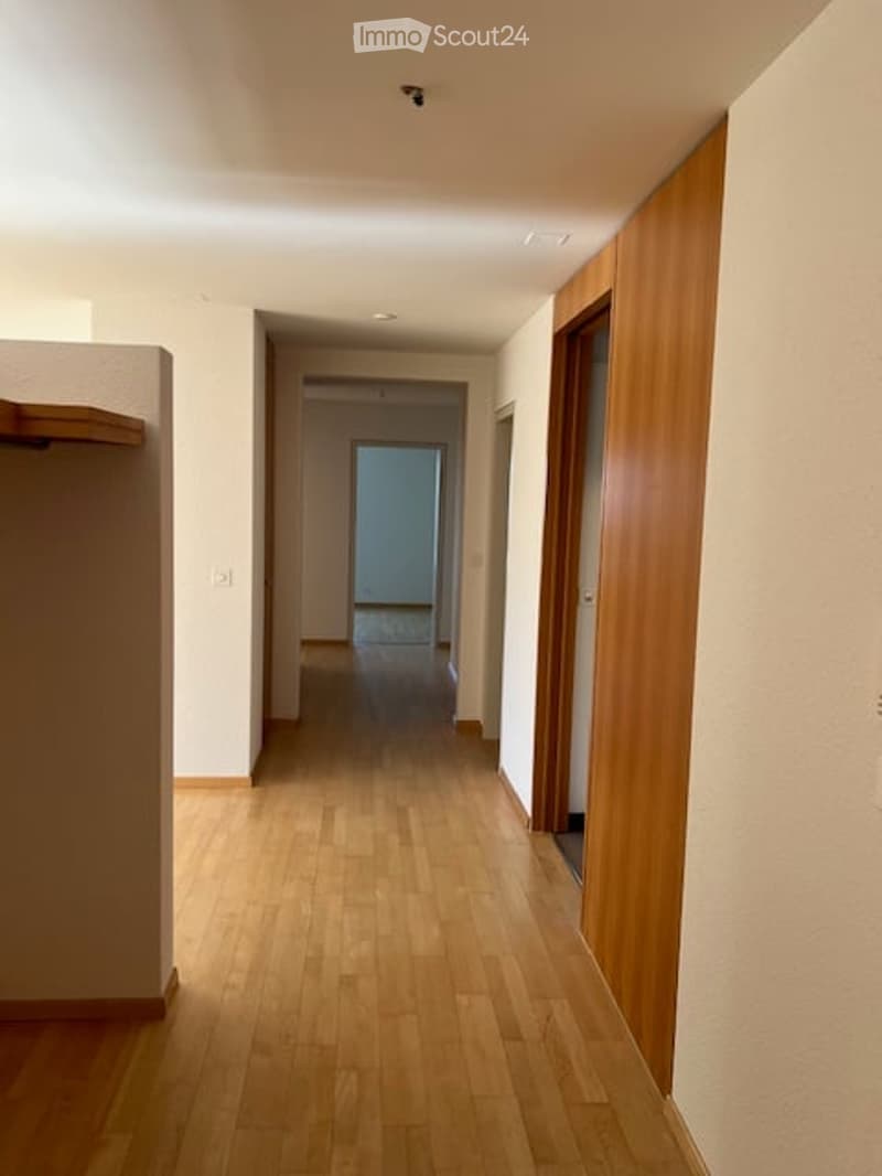 A long, narrow hallway with light wooden flooring and white walls, featuring a wooden door on the right and a small shelf on the left.