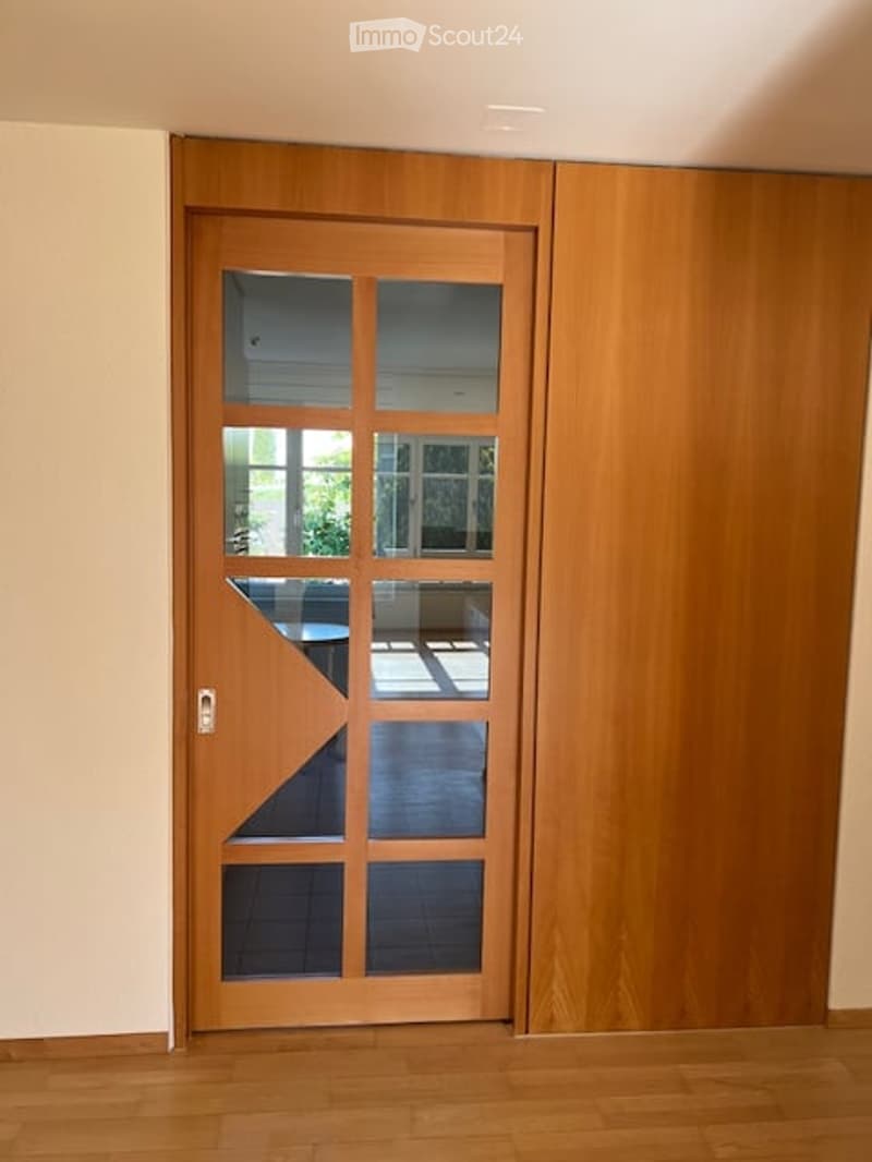 A modern wooden door with glass panels is set into a light-colored wall, featuring a sleek design with a visible handle and a small decorative element on the glass.