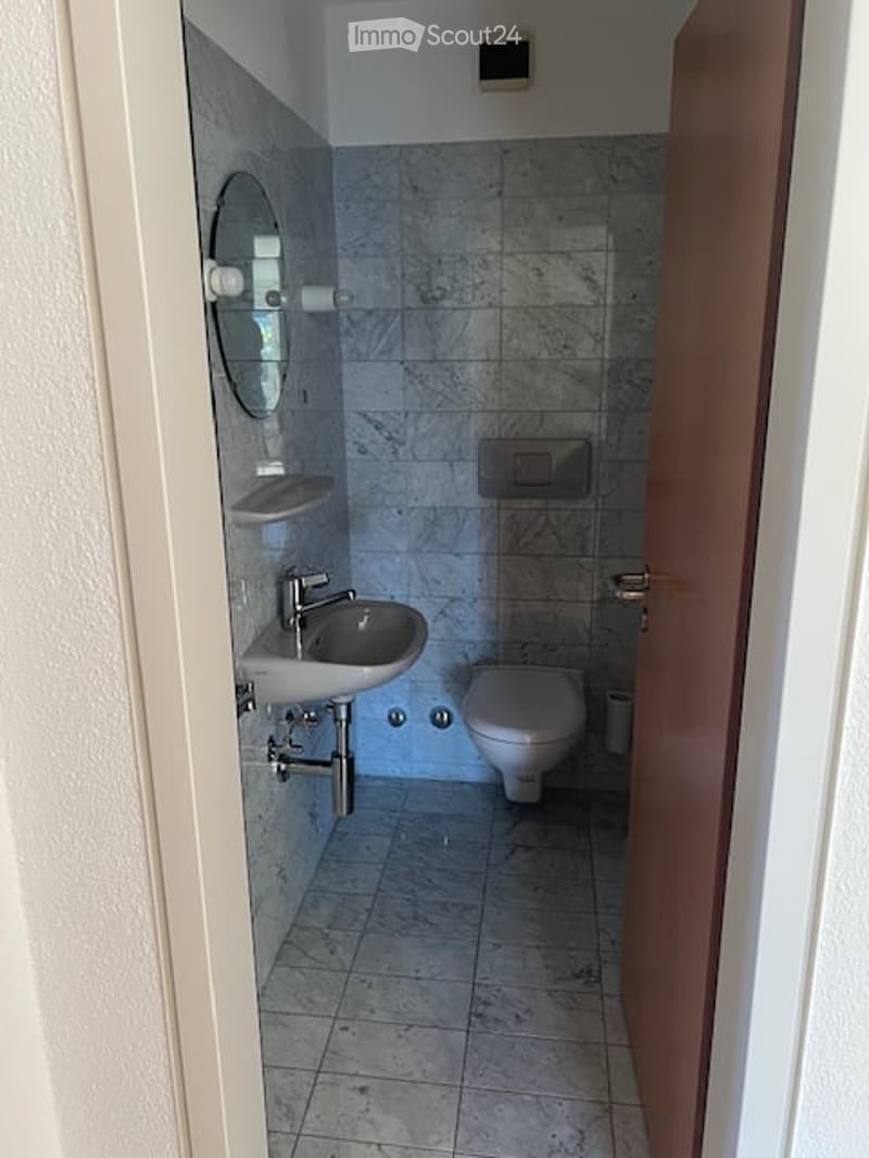 Modern bathroom with grey tiled walls and floor, white fixtures including a sink, faucet, and toilet, a mirror above the sink, and a towel rack on the wall.