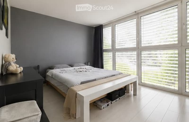 Modern bedroom with large windows, grey walls, a bed with grey bedding, a nightstand, a bench at the foot of the bed, and a teddy bear on a stool.