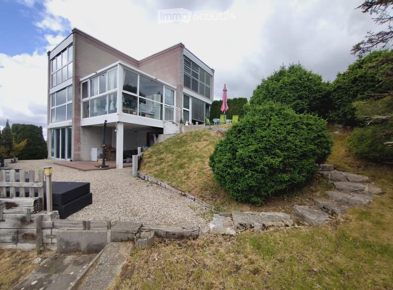 Modern two-story house with large windows and a raised ground floor, surrounded by greenery and a gravel pathway.