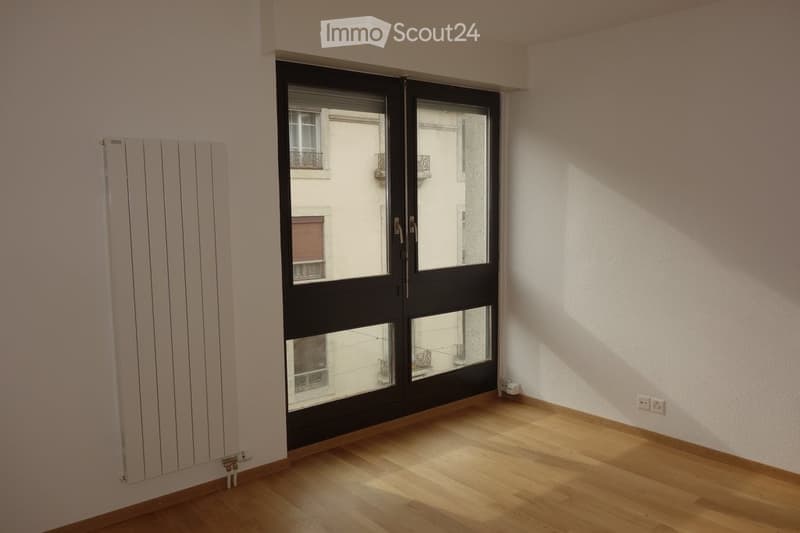 Empty room with wooden flooring, white walls, and large black-framed sliding doors leading to a balcony. A white radiator is mounted on the left wall.