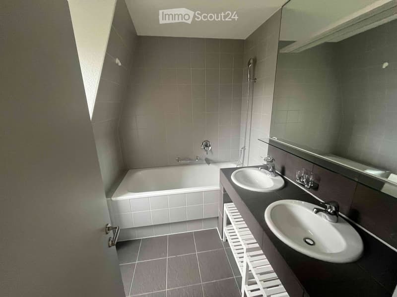 Modern bathroom with a bathtub, two sinks, a large mirror, and grey tiled walls and floor.